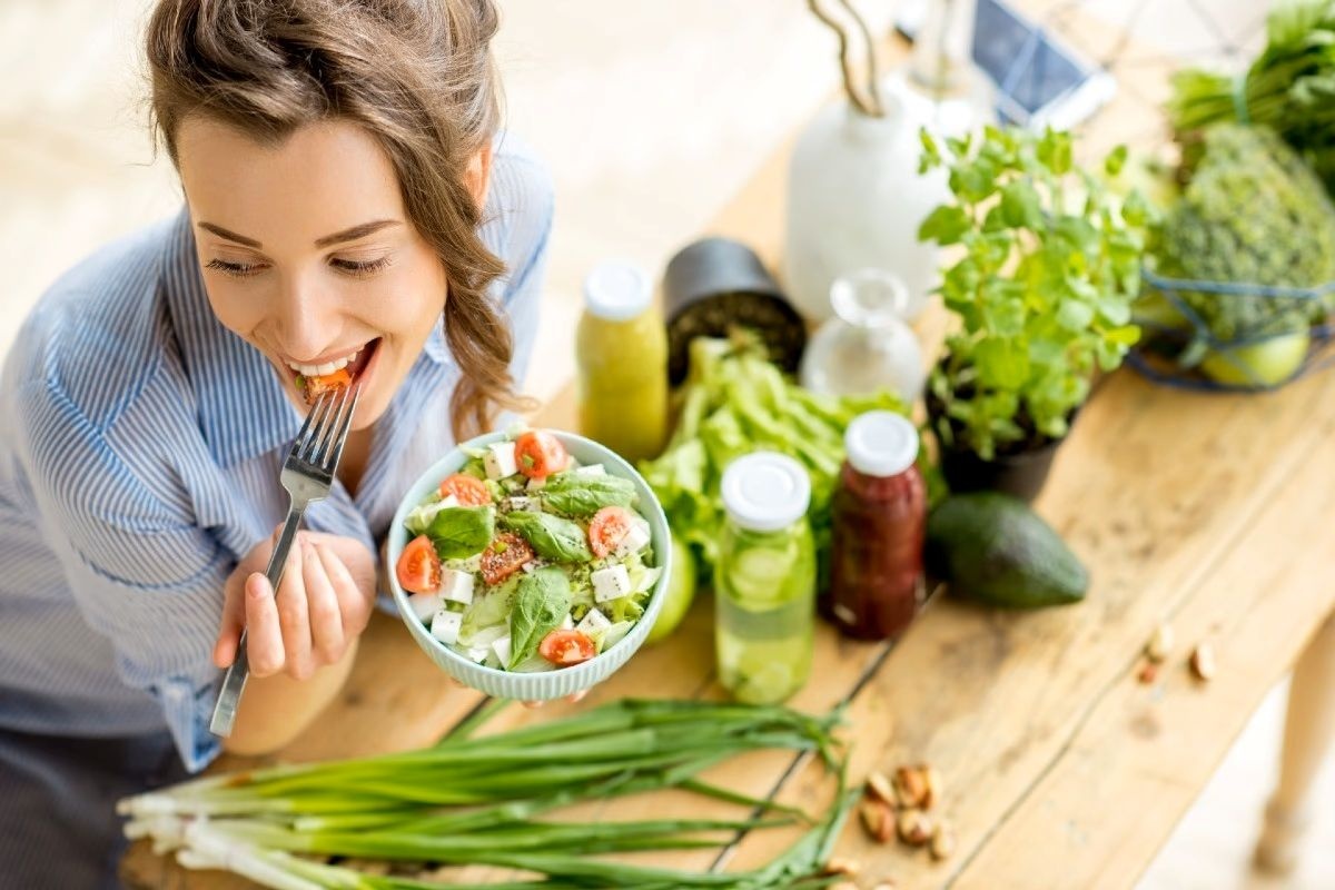 Femeie zâmbitoare care savurează o salată proaspătă lângă băuturi verzi și ingrediente naturale